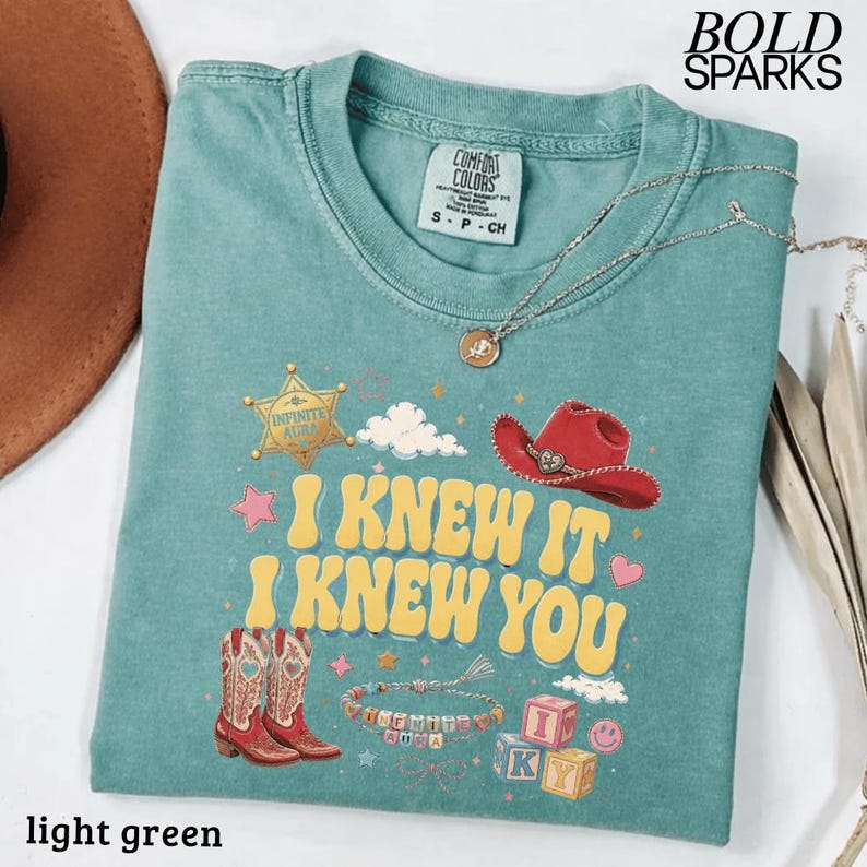 Może przedstawiać: Jasnozielony t-shirt z nadrukiem graficznym z ż&oacute;łtym napisem "I KNEW IT I KNEW YOU". Wz&oacute;r zawiera złotą gwiazdę szeryfa, czerwony kowbojski kapelusz, dwa kowbojskie buty, bransoletkę z koralik&oacute;w oraz klocki z literami. Na koszulce leży srebrny naszyjnik.