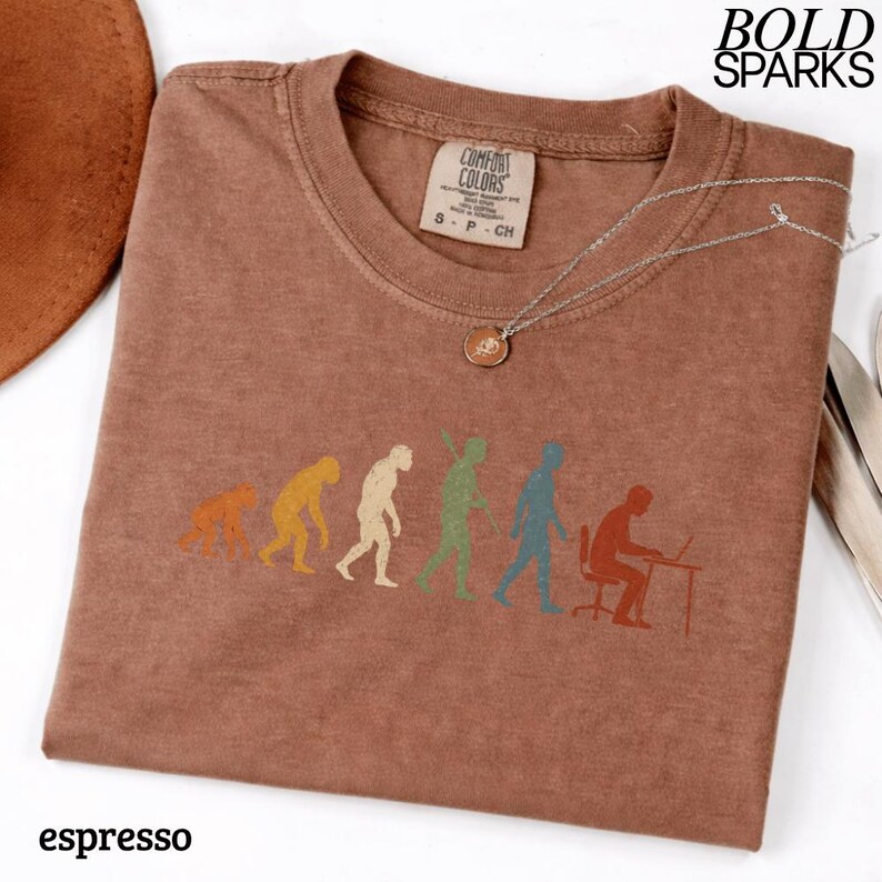 Puede incluir: Una camiseta marr&oacute;n con un estampado gr&aacute;fico de la evoluci&oacute;n humana, desde un primate hasta una persona sentada en un escritorio con un port&aacute;til. La camiseta tiene cuello redondo y una etiqueta de la marca Comfort Colors. El texto BOLD SPARKS es visible.