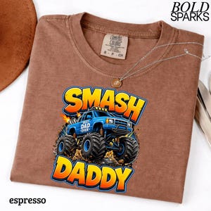 May include: Espresso-colored t-shirt with a graphic of a blue monster truck with the words "SMASH DADDY" in yellow and orange. The shirt also has the words "BEST DAD EVER" on the truck. A silver necklace is draped across the shirt.