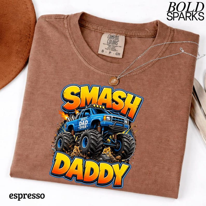 May include: Espresso-colored t-shirt with a graphic of a blue monster truck with the words "SMASH DADDY" in yellow and orange. The shirt also has the words "BEST DAD EVER" on the truck. A silver necklace is draped across the shirt.