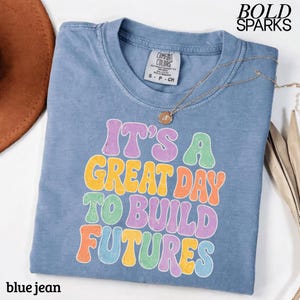 May include: A blue jean Comfort Colors t-shirt with the text "IT'S A GREAT DAY TO BUILD FUTURES" in colorful retro font. A gold necklace and a brown hat are also visible.