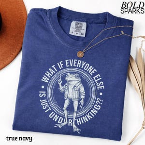 May include: A navy blue short-sleeve t-shirt featuring a white graphic of a frog wearing a jacket. The circular design includes the text "WHAT IF EVERYONE ELSE IS JUST UNDERTHINKING??". The shirt has a Comfort Colors brand label at the neck.