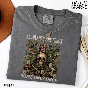 May include: A gray t-shirt with the text "ALL PLANTS ARE EDIBLE SOME ONLY ONCE" and a graphic of a skull surrounded by plants and mushrooms. A gold necklace is draped across the shirt.