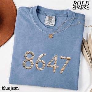 May include: A blue jean Comfort Colors t-shirt with the number 8647 made of small stones. The shirt has a round neckline and a small tag. A gold necklace with a pendant is on the shirt. The words "BOLD SPARKS" are in the upper right corner.