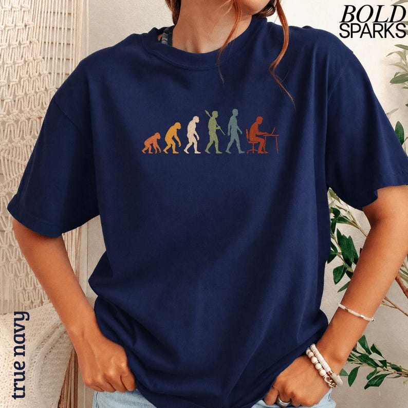 Puede incluir: Una camiseta azul marino de manga corta con un estampado gr&aacute;fico multicolor de la evoluci&oacute;n humana, que comienza con un primate y termina con una persona sentada en un escritorio con un ordenador. El texto BOLD SPARKS aparece en la esquina superior.