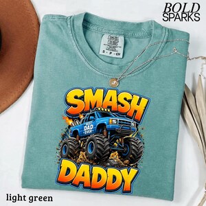 May include: Light green t-shirt with a graphic of a blue monster truck with the words "SMASH DADDY" in yellow and orange. The shirt also has the text "BEST DAD EVER" on the truck. The shirt is made of Comfort Colors material.