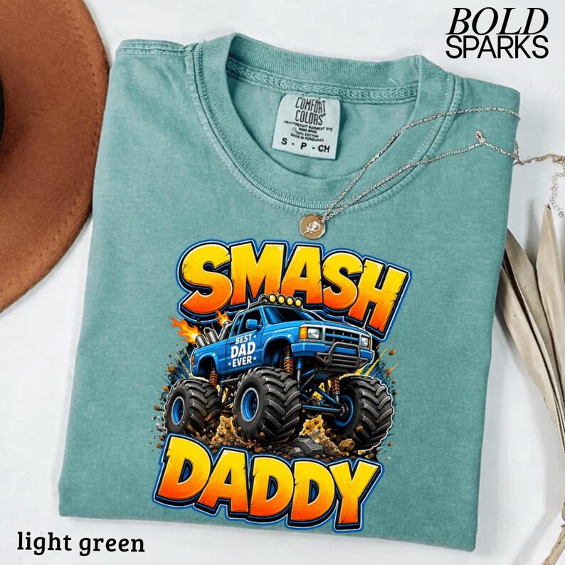 May include: Light green t-shirt with a graphic of a blue monster truck with the words "SMASH DADDY" in yellow and orange. The shirt also has the text "BEST DAD EVER" on the truck. The shirt is made of Comfort Colors material.