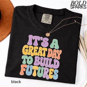 May include: Black t-shirt with the text "IT'S A GREAT DAY TO BUILD FUTURES" in colorful bubble letters. The shirt has a crew neck and short sleeves. A gold necklace with a pendant is draped across the shirt. The word "black" is printed at the bottom.