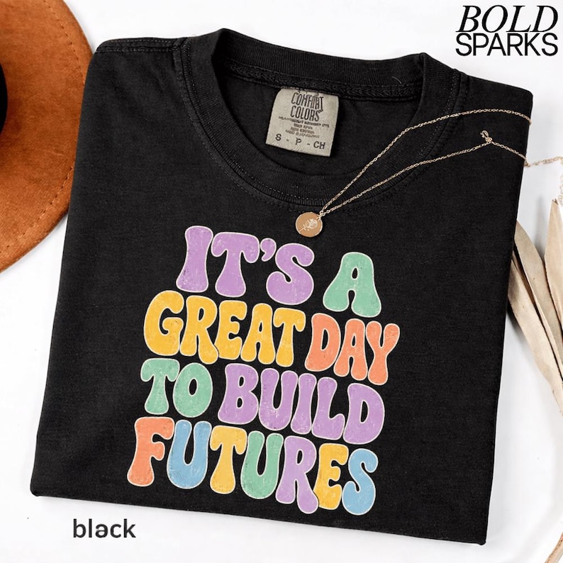 May include: Black t-shirt with the text "IT'S A GREAT DAY TO BUILD FUTURES" in colorful bubble letters. The shirt has a crew neck and short sleeves. A gold necklace with a pendant is draped across the shirt. The word "black" is printed at the bottom.