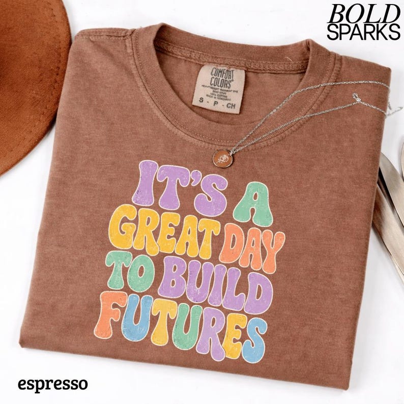 May include: Espresso-colored t-shirt with the text "IT'S A GREAT DAY TO BUILD FUTURES" in colorful, retro-style lettering. The shirt has a crew neck and short sleeves. A silver necklace with a small pendant is draped on the shirt.