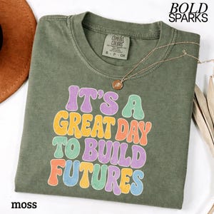 May include: A moss green t-shirt with the text "IT'S A GREAT DAY TO BUILD FUTURES" in colorful bubble letters. A gold necklace with a pendant is draped on the shirt. The brand name "BOLD SPARKS" is in the upper right corner.