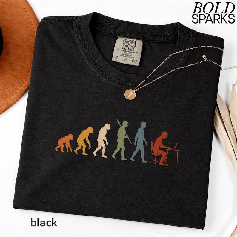 Puede incluir: Una camiseta negra con un estampado gr&aacute;fico de la evoluci&oacute;n humana, desde un primate hasta una persona sentada en un escritorio con un port&aacute;til. La camiseta tiene una etiqueta de la marca Comfort Colors en el cuello y el texto BOLD SPARKS arriba.