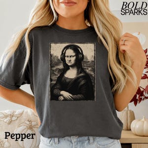 Może przedstawiać: Ciemnoszary t-shirt z nadrukiem graficznym przedstawiającym Monę Lisę w czarnych słuchawkach. Grafika umieszczona jest w prostokątnej ramce i utrzymana w sepiowej, szkicowej stylistyce. W prawym górnym rogu znajduje się napis BOLD SPARKS.