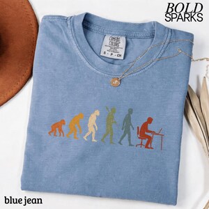 Puede incluir: Una camiseta de color azul jean con un estampado gr&aacute;fico de la evoluci&oacute;n humana, desde un primate hasta una persona sentada en un escritorio con un port&aacute;til. La camiseta tiene cuello redondo y una etiqueta de la marca Comfort Colors en el interior. El texto BOLD SPARKS es visible en la esquina superior derecha.