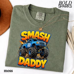 May include: Moss green t-shirt with a graphic of a blue monster truck with the words "SMASH DADDY" in yellow and orange. The shirt also has the text "BEST DAD EVER". A gold necklace with a pendant is on the shirt.