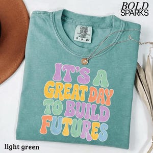 May include: Light green t-shirt with the text "IT'S A GREAT DAY TO BUILD FUTURES" in colorful bubble letters. The shirt has a crew neck and short sleeves. A silver necklace with a pendant is draped on the shirt.
