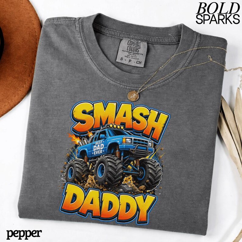 May include: A gray t-shirt with a graphic of a blue monster truck with the words "SMASH DADDY" in yellow and orange. The shirt also has the text "BEST DAD EVER". A gold necklace is draped across the shirt.