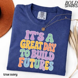 May include: A true navy blue t-shirt with the text "IT'S A GREAT DAY TO BUILD FUTURES" in colorful bubble letters. A gold necklace with a pendant rests on the shirt. The shirt is labeled "Comfort Colors" and is a size small.
