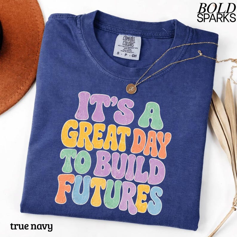 May include: A true navy blue t-shirt with the text "IT'S A GREAT DAY TO BUILD FUTURES" in colorful bubble letters. A gold necklace with a pendant rests on the shirt. The shirt is labeled "Comfort Colors" and is a size small.