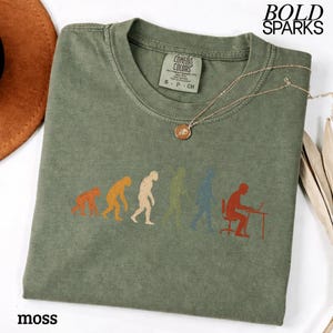 Puede incluir: Una camiseta de cuello redondo color verde musgo con un estampado gr&aacute;fico de la evoluci&oacute;n humana, desde un primate hasta una persona sentada en un escritorio con un port&aacute;til. La prenda tiene la etiqueta Comfort Colors y el texto BOLD SPARKS.