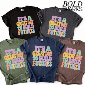 May include: Five different colored t-shirts are displayed, each with the text "IT'S A GREAT DAY TO BUILD FUTURES" in a colorful, retro font. The shirts are black, grey, olive green, navy blue, and brown.