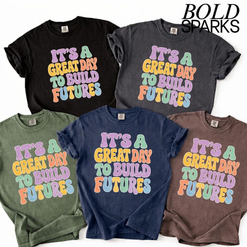May include: Five different colored t-shirts are displayed, each with the text "IT'S A GREAT DAY TO BUILD FUTURES" in a colorful, retro font. The shirts are black, grey, olive green, navy blue, and brown.