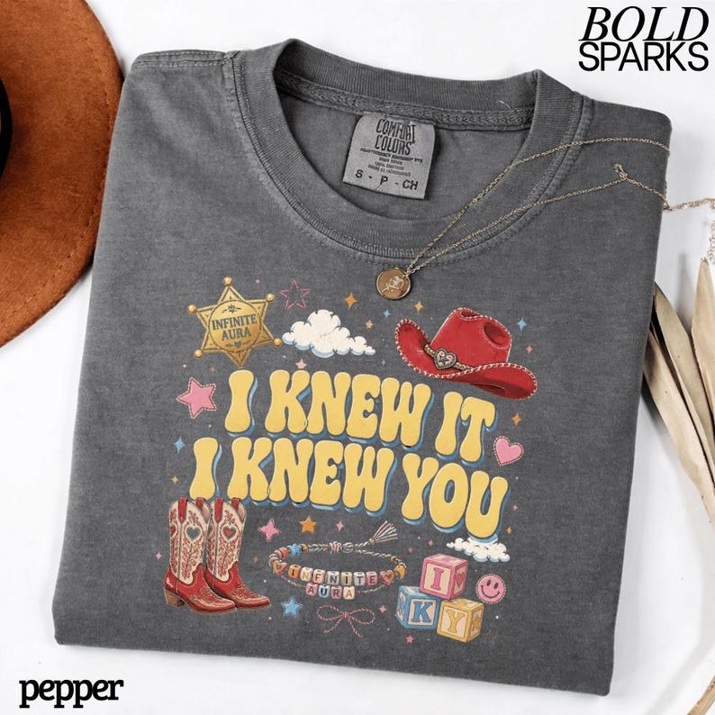Może przedstawiać: Ciemnoszary t-shirt z nadrukiem graficznym z ż&oacute;łtym napisem I KNEW IT I KNEW YOU. Wz&oacute;r zawiera złotą gwiazdę szeryfa, czerwony kowbojski kapelusz, parę kowbojek, bransoletkę z koralik&oacute;w oraz klocki z literami.