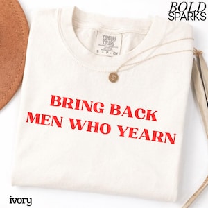 Op de afbeelding: Een ivoorkleurig t-shirt met de rode tekst BRING BACK MEN WHO YEARN op de voorzijde. Het shirt heeft een Comfort Colors merklabel in de nek en wordt vergezeld door een dunne goudkleurige ketting met een kleine ronde hanger.