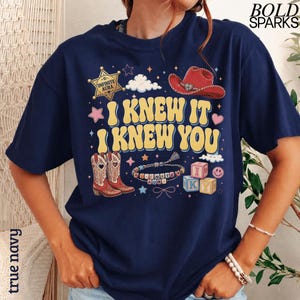Może przedstawiać: Granatowa koszulka z ż&oacute;łtym napisem I KNEW IT I KNEW YOU, otoczonym złotą gwiazdą szeryfa z napisem INFINITE AURA, czerwonym kowbojskim kapeluszem, kowbojkami, bransoletką z koralik&oacute;w, klockami z literami IKY oraz gwiazdkami.
