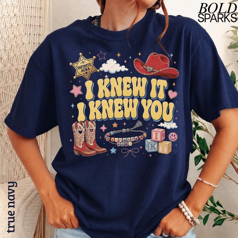 Może przedstawiać: Granatowa koszulka z ż&oacute;łtym napisem I KNEW IT I KNEW YOU, otoczonym złotą gwiazdą szeryfa z napisem INFINITE AURA, czerwonym kowbojskim kapeluszem, kowbojkami, bransoletką z koralik&oacute;w, klockami z literami IKY oraz gwiazdkami.