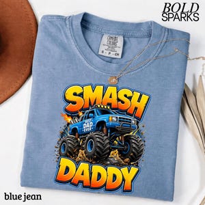 May include: A blue jean colored t-shirt with a graphic of a monster truck that says "SMASH DADDY". The monster truck is blue with the words "BEST DAD EVER" on the side. The shirt has a crew neck and short sleeves.
