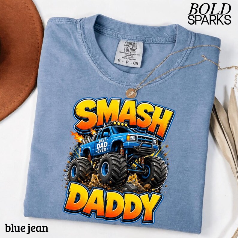 May include: A blue jean colored t-shirt with a graphic of a monster truck that says "SMASH DADDY". The monster truck is blue with the words "BEST DAD EVER" on the side. The shirt has a crew neck and short sleeves.