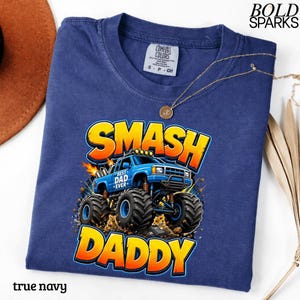 May include: A true navy blue t-shirt featuring a graphic of a blue monster truck with the words "SMASH DADDY" in yellow and orange. The shirt also has the text "BEST DAD EVER" on the truck. A gold necklace is draped across the shirt.