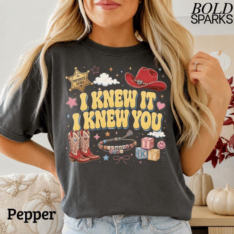 Może przedstawiać: Ciemnoszary t-shirt z nadrukiem graficznym z ż&oacute;łtym napisem "I KNEW IT I KNEW YOU". Wz&oacute;r zawiera złotą gwiazdę szeryfa, czerwony kowbojski kapelusz, dwa kowbojskie buty, bransoletkę z koralik&oacute;w oraz klocki z literami tworzące napis "I KY".
