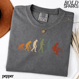 Puede incluir: Una camiseta gris carb&oacute;n de manga corta con un estampado gr&aacute;fico de la evoluci&oacute;n humana, desde un primate hasta una persona sentada en un escritorio con un ordenador. La camiseta tiene cuello redondo y una etiqueta de la marca Comfort Colors.