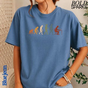 Puede incluir: Una camiseta azul de manga corta con un estampado gr&aacute;fico de la evoluci&oacute;n humana, que progresa desde un primate hasta una persona sentada en un escritorio con un ordenador. El texto BOLD SPARKS aparece en la esquina superior derecha.
