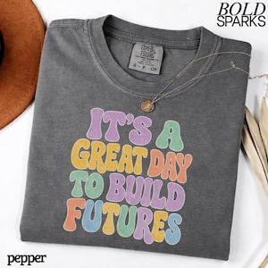 May include: A charcoal gray t-shirt with the text "IT'S A GREAT DAY TO BUILD FUTURES" in colorful retro font. The shirt has a crew neck and short sleeves. A gold necklace with a pendant is draped on the shirt. The brand name "COMFORT COLORS" is on the tag.