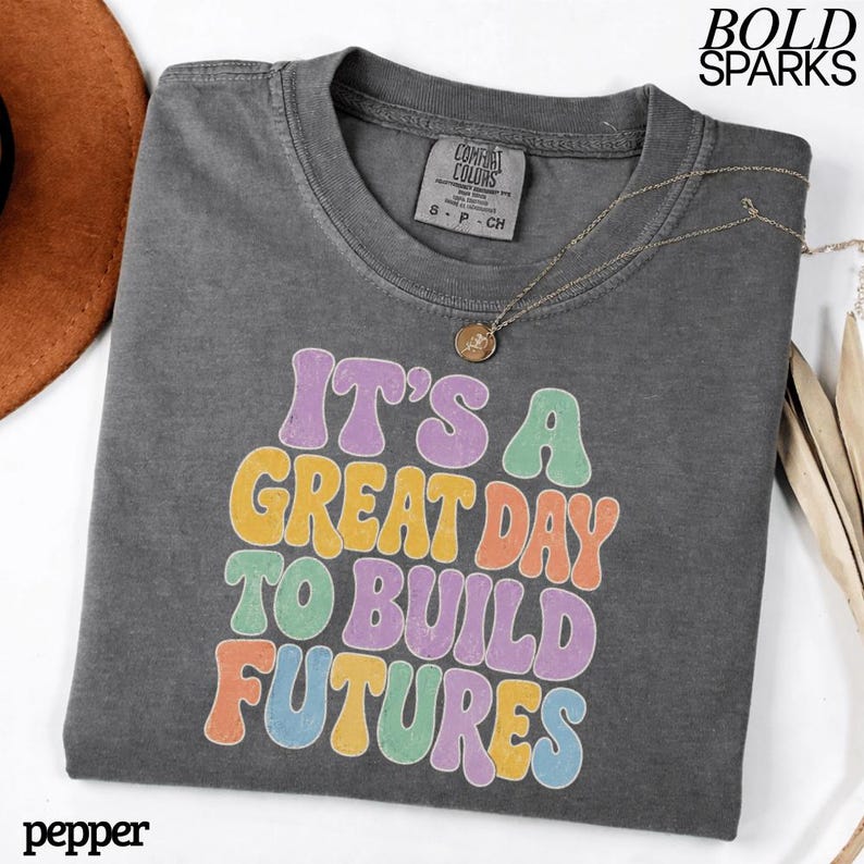 May include: A charcoal gray t-shirt with the text "IT'S A GREAT DAY TO BUILD FUTURES" in colorful retro font. The shirt has a crew neck and short sleeves. A gold necklace with a pendant is draped on the shirt. The brand name "COMFORT COLORS" is on the tag.