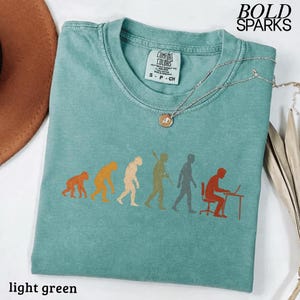 Puede incluir: Una camiseta de manga corta de color verde claro con un estampado gr&aacute;fico de la evoluci&oacute;n humana, desde un primate hasta una persona sentada en un escritorio con un port&aacute;til. La camiseta tiene cuello redondo y una etiqueta Comfort Colors interior.