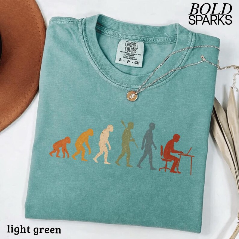 Puede incluir: Una camiseta de manga corta de color verde claro con un estampado gr&aacute;fico de la evoluci&oacute;n humana, desde un primate hasta una persona sentada en un escritorio con un port&aacute;til. La camiseta tiene cuello redondo y una etiqueta Comfort Colors interior.