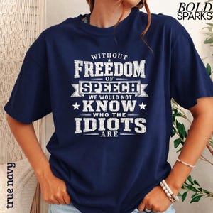 May include: A navy blue short-sleeved t-shirt featuring white distressed text that reads: WITHOUT FREEDOM OF SPEECH WE WOULD NOT KNOW WHO THE IDIOTS ARE. The design includes decorative stars and horizontal lines framing the central text block.