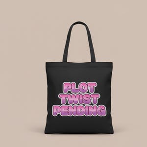 May include: Black tote bag with the text "PLOT TWIST PENDING" in a bubble font with pink and purple gradient colors. The bag has black handles and is made of a sturdy material, suitable for carrying various items.