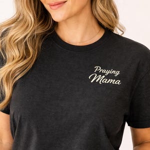 May include: Dark grey t-shirt with the text "Praying Mama" in white script. The shirt has short sleeves and a crew neck. The model is wearing the shirt.