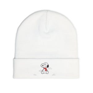 May include: A white beanie featuring an embroidered Snoopy design. Snoopy is wearing a red scarf and is sitting down. The beanie is made of a soft, knitted material.
