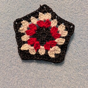 May include: A black, five-sided crochet piece with a floral design. The flower petals are red and white, and the center is black. The piece is handmade and sits on a light blue surface.