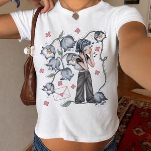 May include: White crop top featuring a whimsical illustration of a person wearing headphones, surrounded by bluebell flowers and musical notes. A pink envelope is also in the design. The person in the illustration is wearing a black top and gray pants.
