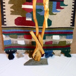 May include: A wooden sculpture made from twisted branches, standing on a slate base. The sculpture is in front of a colourful woven rug with a geometric design.