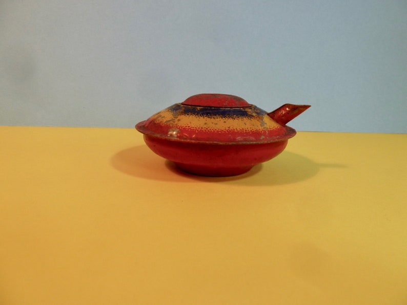 Puede incluir: Una tetera de metal roja, amarilla y azul con un pico y una tapa. La tetera est&aacute; ligeramente desgastada y tiene un acabado moteado.