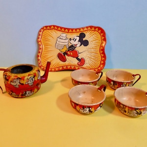 May include: Vintage Mickey Mouse tin tea set with a red teapot, four cups, and a tray featuring Mickey Mouse carrying a stack of dishes. The set is decorated with yellow, red, and black details.
