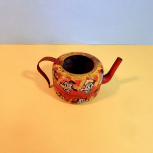 May include: A small, red and yellow metal teapot with a cartoon character design. The teapot has a spout and a handle.